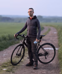 Self‑portrait of a cyclist on a field track after sunset, using a phone to trigger the shot.