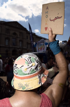 Participant at a Paris LGBT demonstration, arm raised with a cardboard sign reading “Amour” and the Arabic word for love, in front of a Louvre-side street.