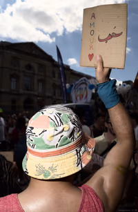 Participant at a Paris LGBT demonstration, arm raised with a cardboard sign reading “Amour” and the Arabic word for love, in front of a Louvre-side street.