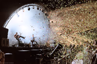 High-kicking guitarist and bandmate under an arched truss as confetti storms from the right; Mons station’s curves glow in the background.