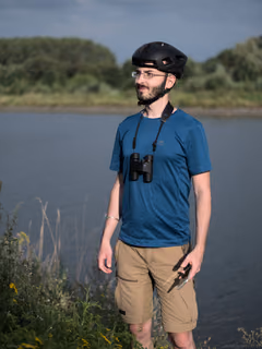 Riverside self‑portrait with bicycle helmet, pocket binoculars, and phone remote; flat, even lighting.